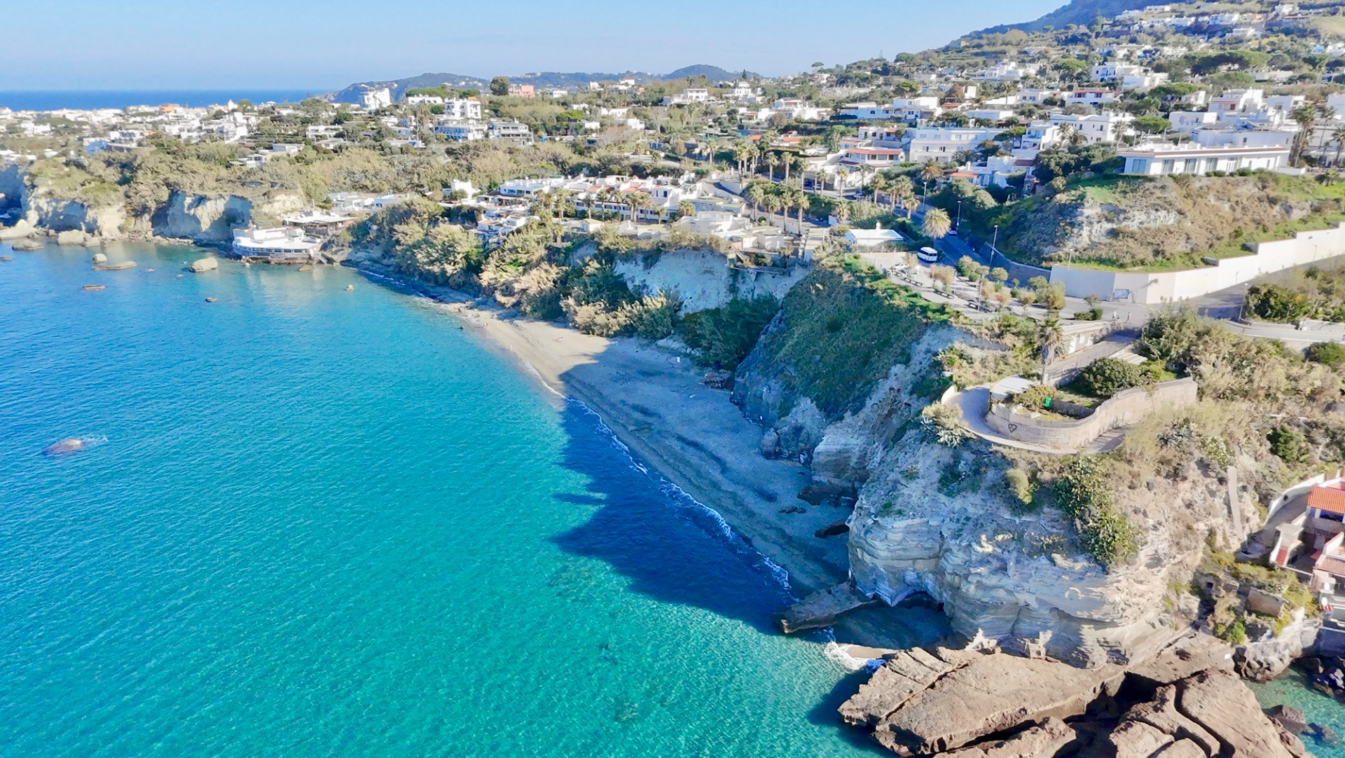 Aerial view of a coastal town with white buildings, turquoise water, and rocky shoreline.
