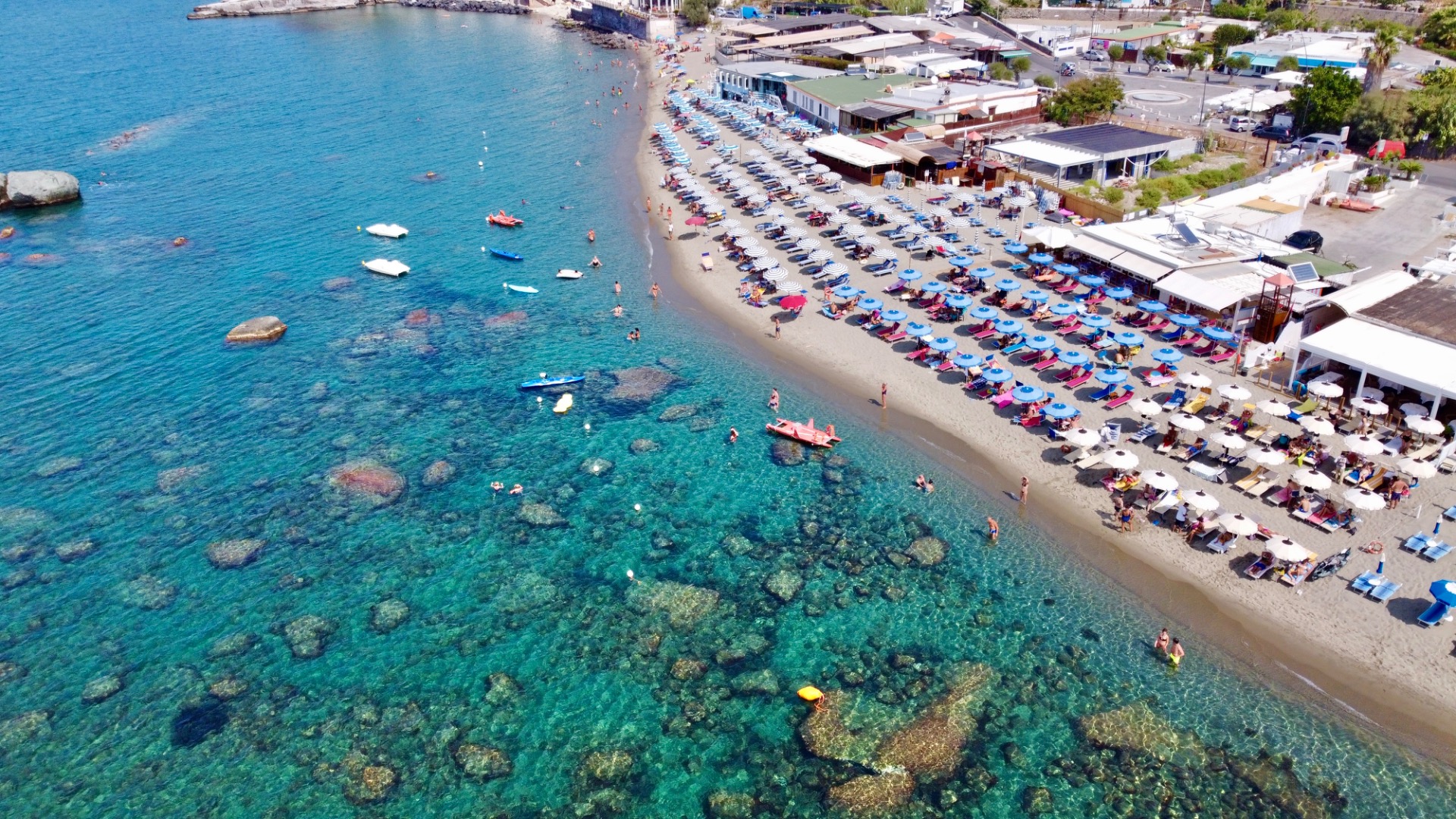 Aerial view of a crowded beach with rows of umbrellas, clear turquoise water, and boats.