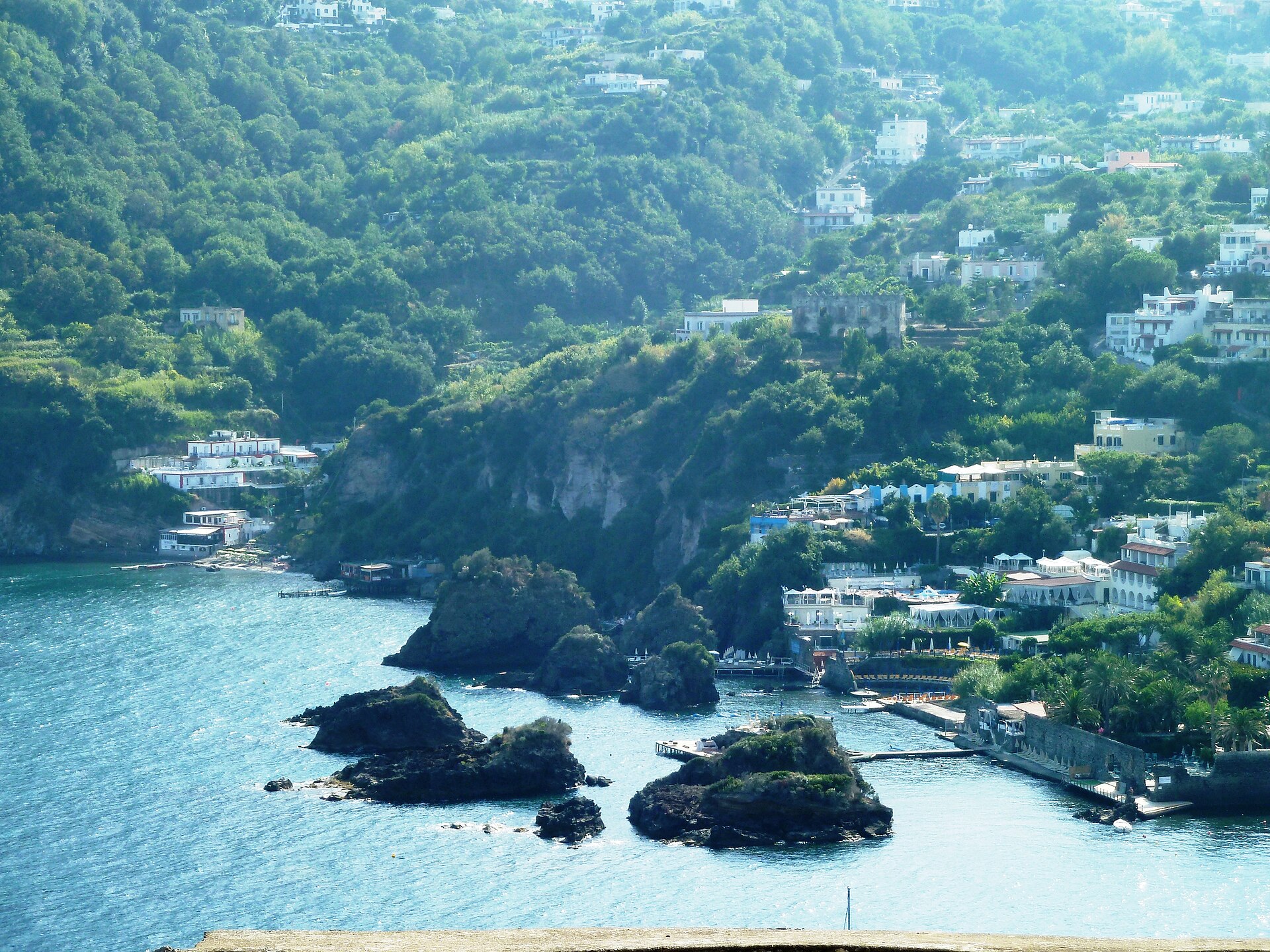 Coastal hillside town with white buildings, lush green trees, and rocky islands in blue water.