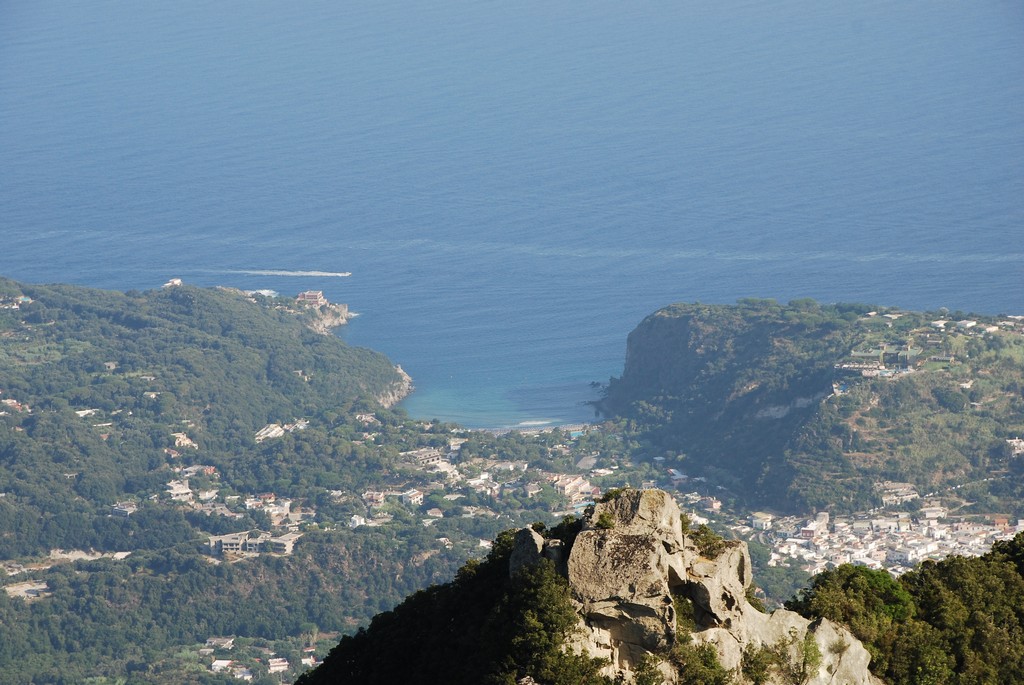 Vista panoramica di una costa mediterranea con scogli, villaggi e mare blu tra colline verdi.