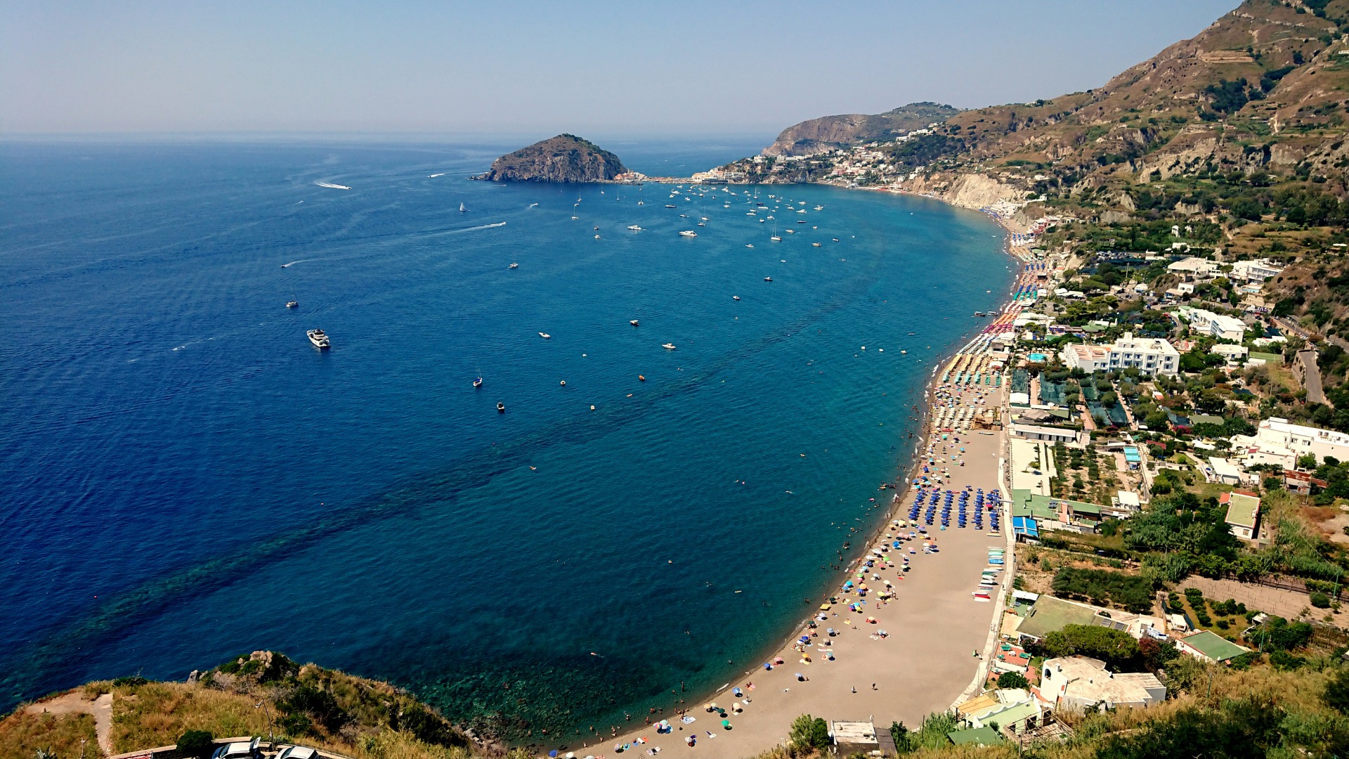 Baia costiera con spiaggia affollata, barche e yacht in mare, scogliere verdi e case sulla costa.
