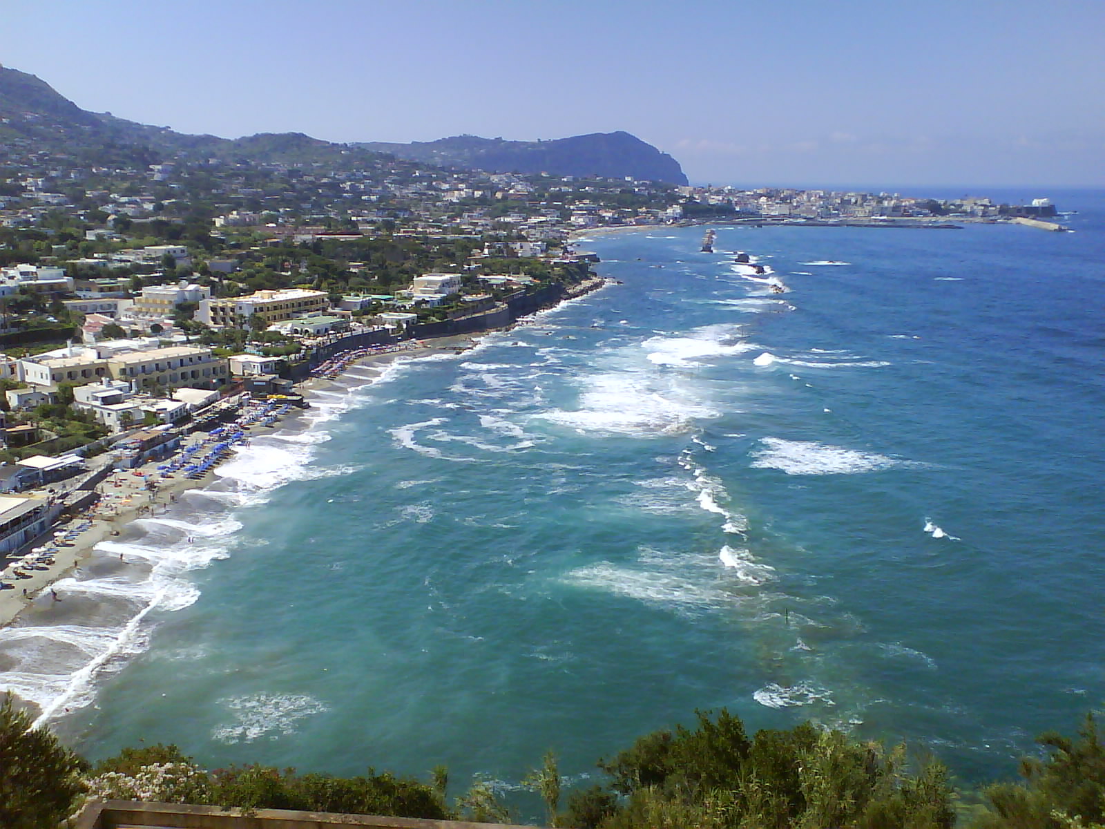 Veduta costiera con spiaggia affollata, onde e case bianche lungo una baia ai piedi di una collina.