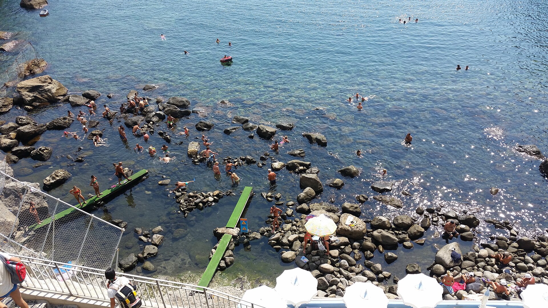 Bagnanti nuotano e si tuffano in mare limpido tra rocce, con gommoni e scivoli verdi.