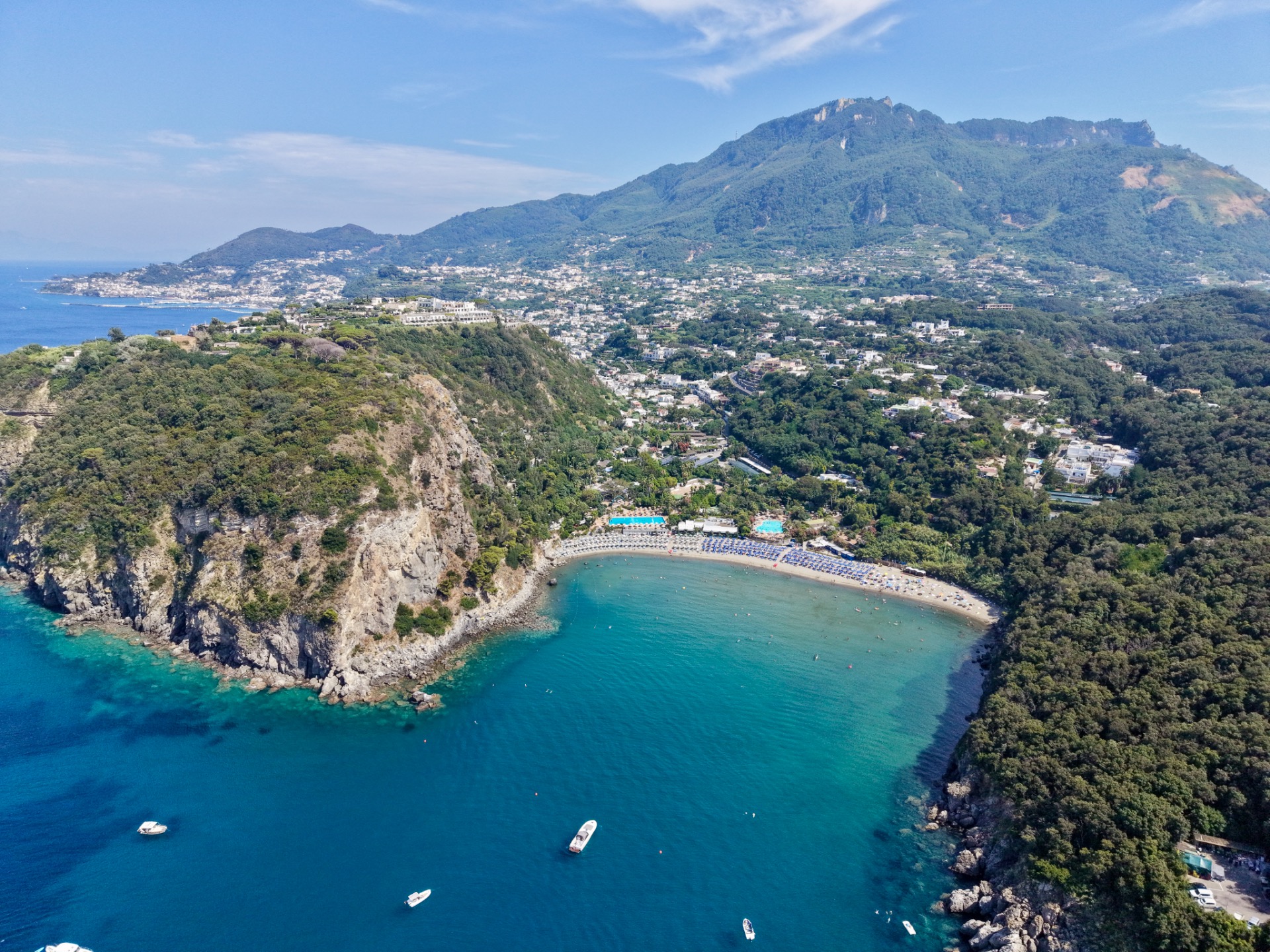 Vista panoramica di una costa mediterranea con scogli, villaggi e mare blu tra colline verdi.