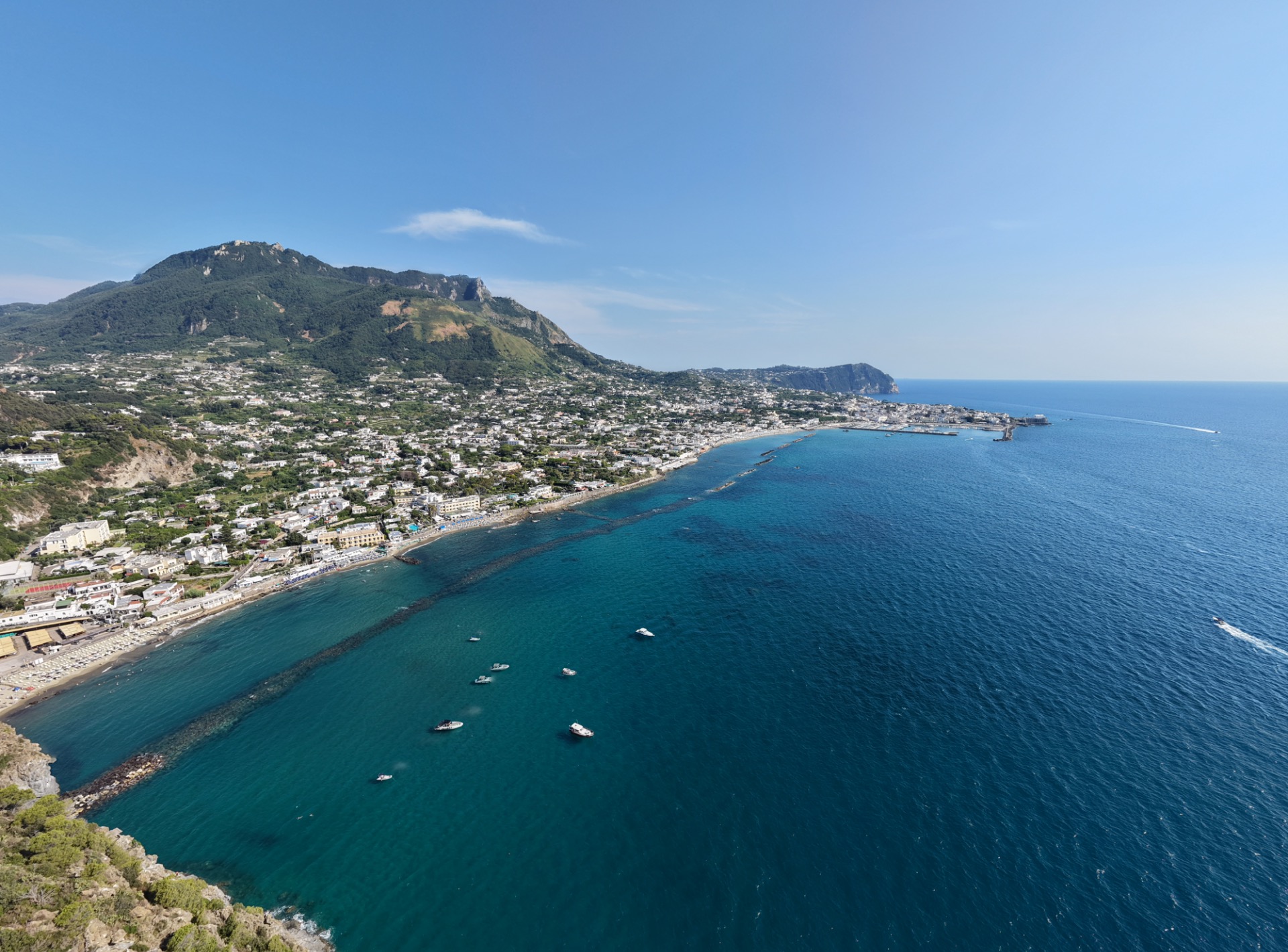 Veduta costiera con spiaggia affollata, onde e case bianche lungo una baia ai piedi di una collina.