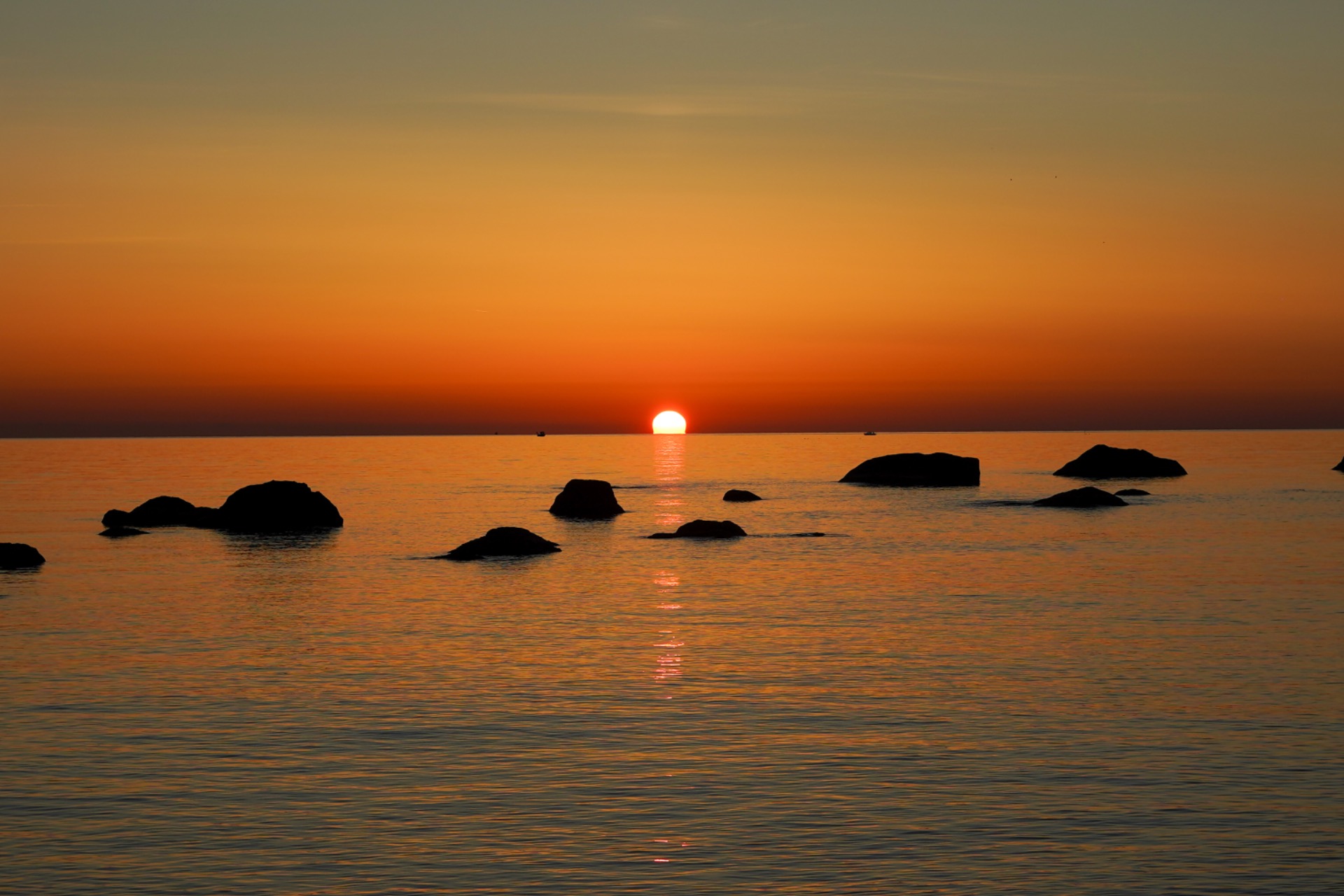 Tramonto sul mare arancione con sole basso, rocce in primo piano e riflessi sull’acqua calma.