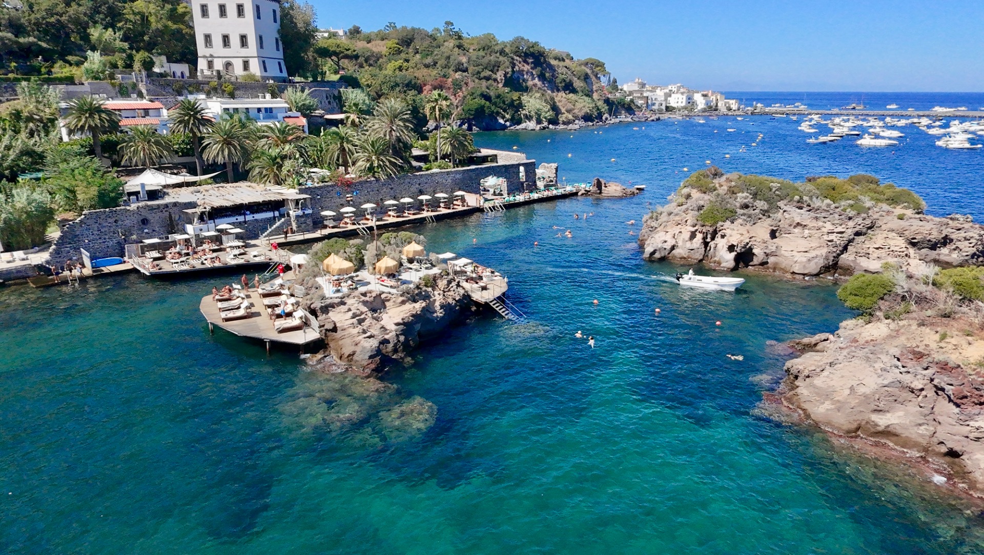 Coastal hillside town with white buildings, lush green trees, and rocky islands in blue water.