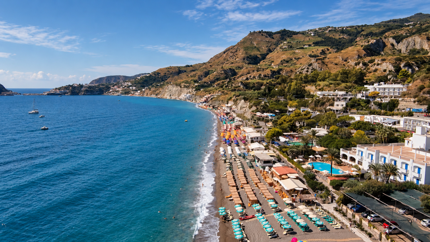 Aerial view of a coastal bay with sailboats, cliffs, and a crowded sandy beach.